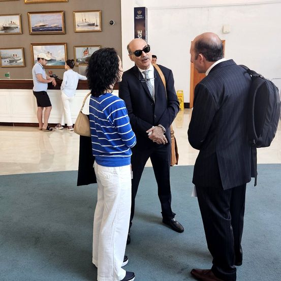 Drei Menschen stehen und unterhalten sich in einer Lobby. Eine Frau in weißen Hosen schaut einen Mann mit Sonnenbrille an. Ein anderer Mann trägt einen Rucksack.