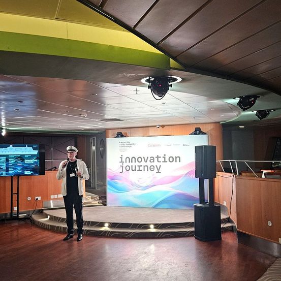Ein Mann steht auf der Bühne, hält ein Mikrofon in der Hand, mit einem großen Banner hinter ihm, das 'Innovation Journey' steht. Es gibt Lautsprecher und einen Monitor auf der linken Seite, und die Bühne ist gut beleuchtet.