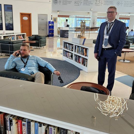 Zwei Männer in einer Bibliothek. Einer sitzt auf einem Stuhl und spricht am Handy. Der andere steht lächelnd, beide sind formell gekleidet. Hinter ihnen sind Bücherregale und Stühle zu sehen.