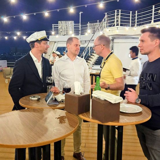Vier Männer stehen um einen Tisch auf dem Deck eines Kreuzfahrtschiffs unter Lichterketten. Einer trägt einen marineblauen Anzug, ein anderer ein weißes Hemd und die anderen zwei gelbe und schwarze Hemden.