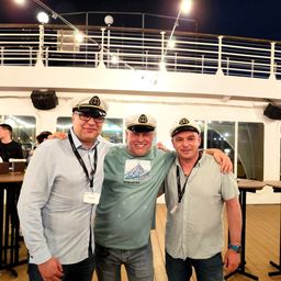 Drei Männer stehen auf dem Deck eines Kreuzfahrtschiffs, lächeln und posieren für ein Foto. Ein Mann trägt ein blaues Hemd, während die anderen grüne Hemden und Kapitänsmützen tragen.