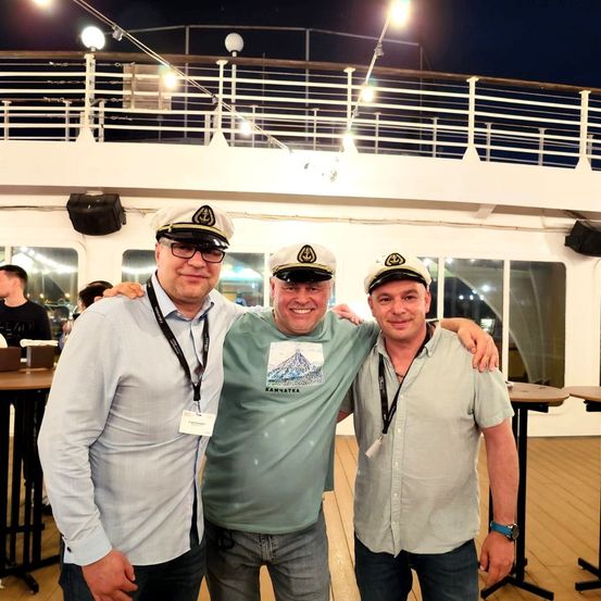 Drei Männer stehen auf dem Deck eines Kreuzfahrtschiffs, lächeln und posieren für ein Foto. Ein Mann trägt ein blaues Hemd, während die anderen grüne Hemden und Kapitänsmützen tragen.