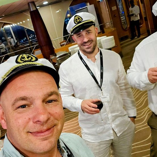 Drei Besatzungsmitglieder sind auf einem Schiff. Der Mann vorne lächelt und trägt eine Kappe. Der Mann in der Mitte hält ein Weinglas und trägt ein weißes Hemd und einen schwarzen Ausweis. Ein anderer Mann ist rechts und trägt ein weißes Hemd und einen Hut.