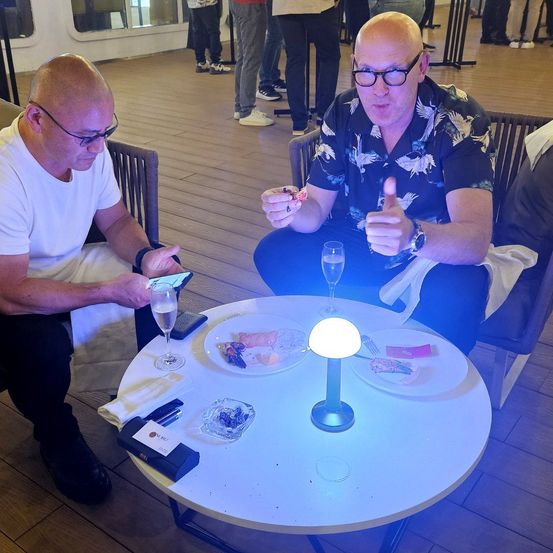 Zwei Männer sitzen an einem Tisch, einer benutzt ein Smartphone und der andere gibt einen Daumen hoch. Sie haben Teller mit Essen und eine Lampe auf dem Tisch. Im Hintergrund sind andere Leute.
