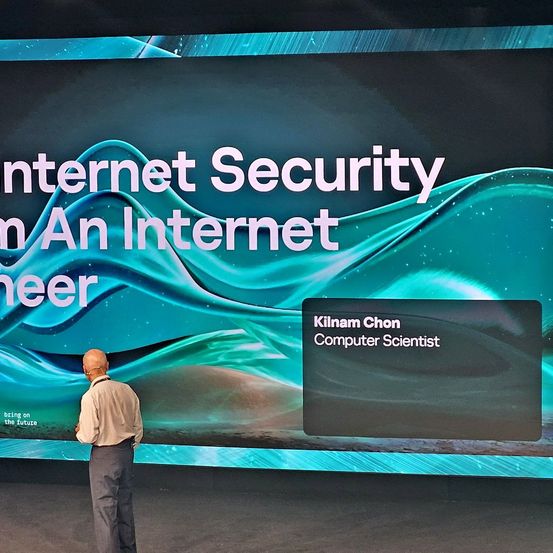 Ein Mann steht vor einem großen digitalen Banner, das über Internetsicherheit spricht. Das Banner zeigt den Namen Kilnam Chon, einem Computerwissenschaftler, vor einem Hintergrund von Wellen.