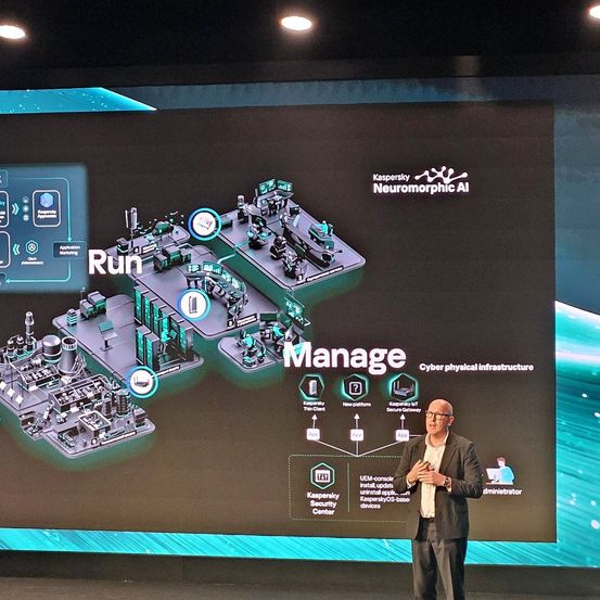 Ein Mann hält eine Präsentation vor einer großen Leinwand, die ein Diagramm mit den Worten 'Laufen' und 'Verwalten' sowie 'Cyber-Physische Infrastruktur' zeigt. Er trägt einen Anzug und eine Brille.