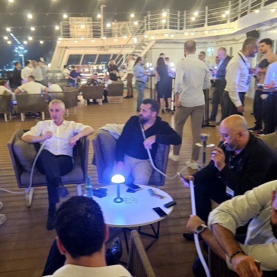 Eine Gruppe von Männern sitzt um einen Tisch auf einer Terrasse herum, benutzt Wasserpfeifen und eine Tischlampe. Der Bereich ist gut beleuchtet mit Lichterketten.