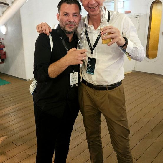 Zwei Männer stehen auf dem Deck eines Schiffes, einer hält ein Bier und der andere eine Flasche, beide lächeln.