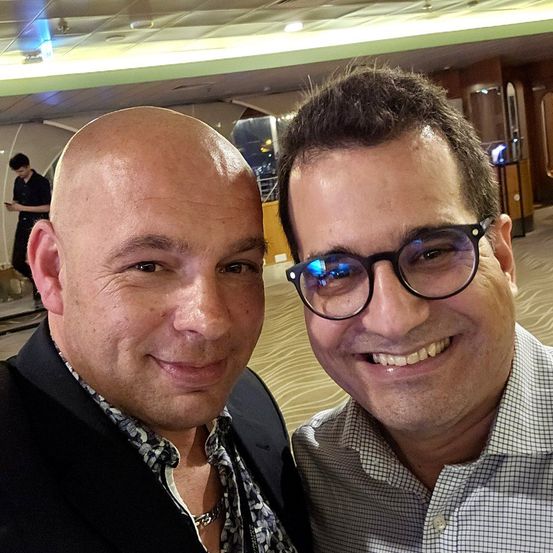 Zwei Männer lächeln für ein Selfie. Einer ist kahlköpfig, der andere trägt eine Brille, beide in Business-Kleidung, in einer Innenraumaufnahme.