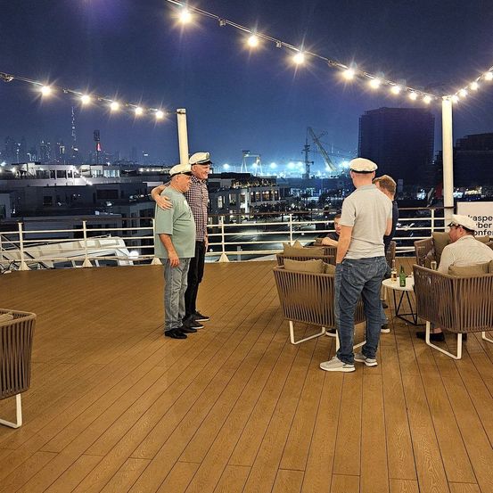 Mehrere Personen stehen bei Nacht auf einer Terrasse mit Blick auf die Stadt. Einige Männer tragen Hüte, und es gibt Stühle und Tische mit Getränken. Lichter hängen an Stangen.