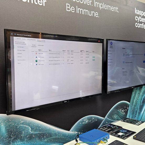 Monitore zeigen eine Kaspersky Sicherheitsgateway-Oberfläche. Im Hintergrund stehen 'Sei Immun' und 'Kaspersky Cyber Conf'. Ein blaues Gehäuse mit Kabeln auf dem Tisch.