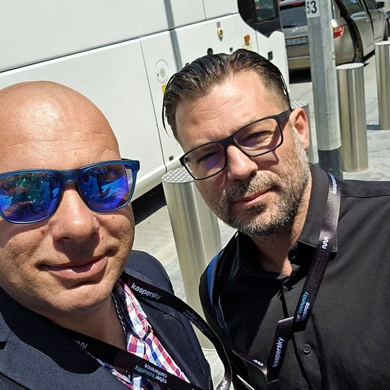 Zwei Männer in Geschäftskleidung stehen neben einem Bus und einem Straßenschild. Einer hat blaue Sonnenbrille und ein Lächeln; der andere hat einen Bart und eine Brille.