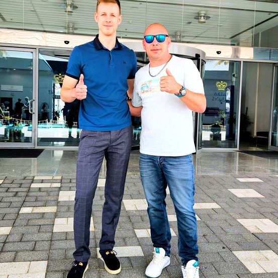 Zwei Männer stehen draußen und geben beide Daumen hoch. Einer trägt ein blaues Hemd und graue Hose, der andere ein weißes Hemd und blaue Jeans. Sie stehen auf einem gepflasterten Boden mit Glastüren hinter ihnen.
