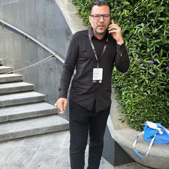 Ein Mann steht auf einer Treppe, hält eine Zigarette, trägt eine Brille und ein schwarzes Hemd und telefoniert.