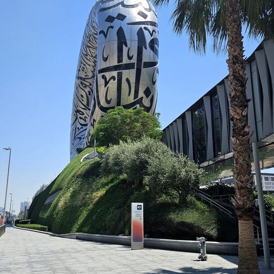 Eine große Skulptur mit arabischen Schriftzeichen steht auf einem grünen Hügel mit Bäumen. Ein Weg mit einem Schild und einem Feuerhydranten befindet sich in der Nähe.
