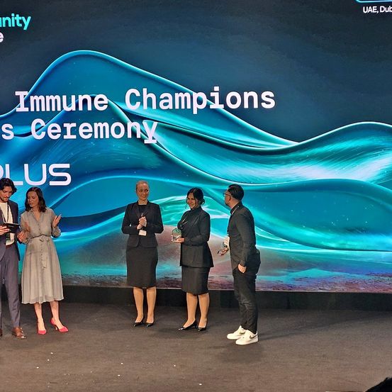 Fünf Personen stehen auf einer Bühne vor einer Kulisse, die 'Immune Champions Ceremony' anzeigt. Die Personen halten Auszeichnungen, und zwei von ihnen sind Frauen. Die Bühne befindet sich unter einem blauen Himmel.