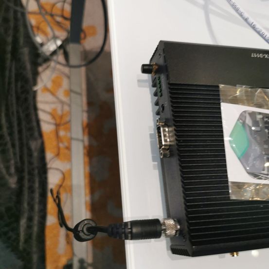 Ein schwarzes elektronisches Gerät mit einer Plastikabdeckung und mehreren Anschlüssen ist an ein Kabel angeschlossen. Es steht auf einer weißen Oberfläche mit verschiedenen Kabeln in der Nähe.