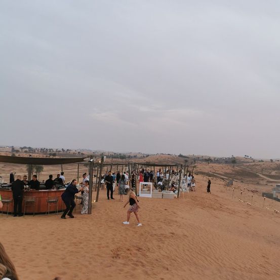 Eine Gruppe von Menschen versammelt sich in einer Wüstenlandschaft für eine Veranstaltung, mit einem Barbereich und Leuten, die in dem Sand herumschlendern.