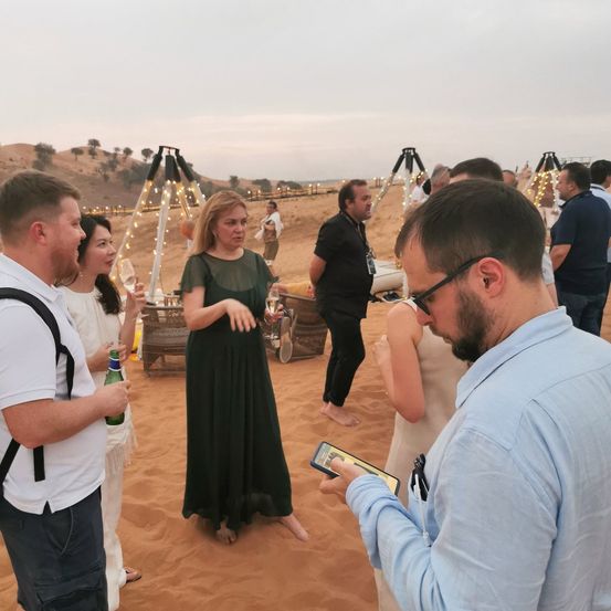 Eine Gruppe von Menschen steht auf einem sandigen Strand, möglicherweise während eines Sonnenuntergangs. Ein Mann in einem blauen Hemd schaut auf sein Handy. Eine Frau in einem schwarzen Kleid steht in der Mitte. Hinter ihnen stehen mehrere Menschen, die sie möglicherweise beobachten. Es gibt einige Objekte auf dem Sand, möglicherweise ein Zelt. In der Ferne gibt es Bäume und Berge.