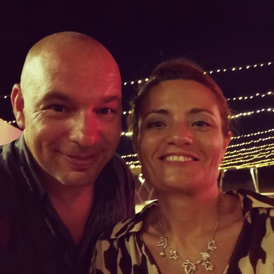 Ein lächelnder Mann und eine Frau stehen bei Nacht eng beieinander, beleuchtet von Lichterketten. Der Mann trägt ein schwarzes Hemd, und die Frau hat eine Halskette.