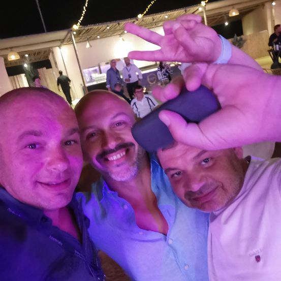 Drei Männer posieren für ein Selfie bei Nacht mit lila Beleuchtung. Der Mann auf der rechten Seite trägt ein weißes Hemd. Im Hintergrund stehen Leute in der Nähe einer Bar.