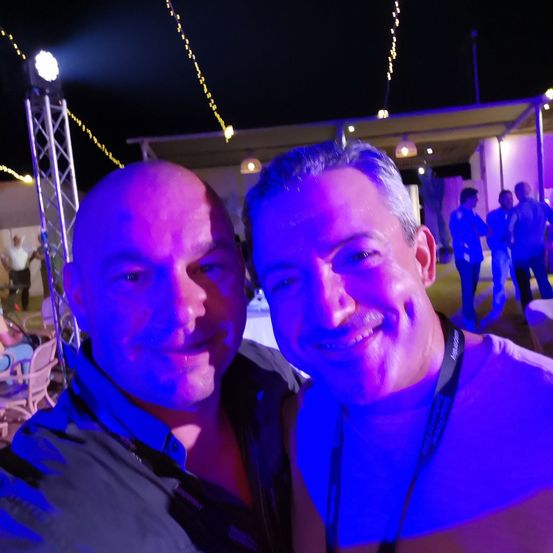 Zwei Männer lächeln für ein Selfie bei einer nächtlichen Veranstaltung, beleuchtet von lila Lichtern, mit unscharfem Hintergrund, der Menschen und Dekoration zeigt.