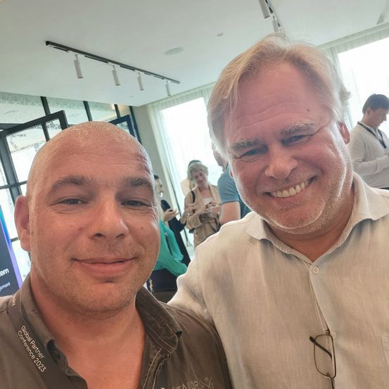 Zwei Männer lächeln in einem Konferenzraum, einer trägt ein Hemd mit der Aufschrift Global Partner Conference 2023. Hinter ihnen stehen und unterhalten sich Leute in der Nähe eines Glasfensters.