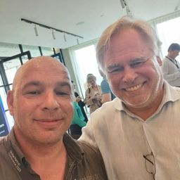 Zwei Männer lächeln in einem Konferenzraum, einer trägt ein Hemd mit der Aufschrift Global Partner Conference 2023. Hinter ihnen stehen und unterhalten sich Leute in der Nähe eines Glasfensters.