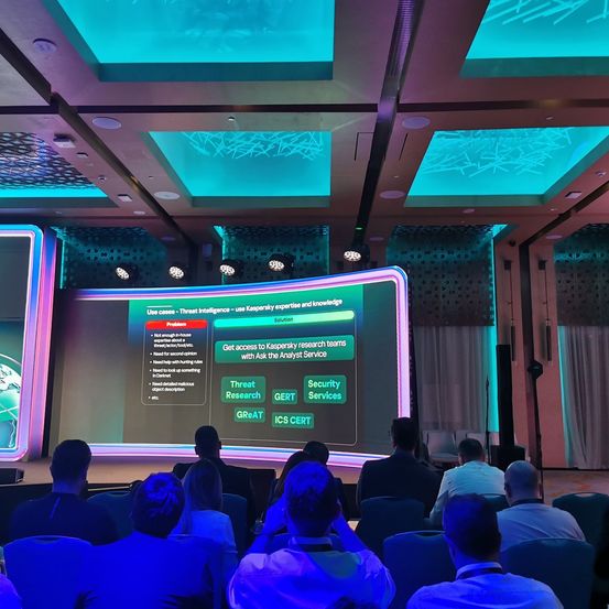 Ein Konferenzsaal mit einer Projektionsleinwand, auf der eine Folie über Bedrohungsaufklärung und Kaspersky zu sehen ist. Das Publikum sitzt auf Stühlen.