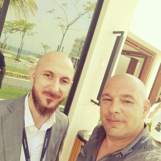 Zwei Männer stehen nah beieinander und lächeln in die Kamera. Einer trägt einen grauen Blazer und Ausweis, der andere ein graues Hemd mit Abzeichen. Hinter ihnen ist eine Glaswand mit Blick auf Bäume und ein Gebäude.