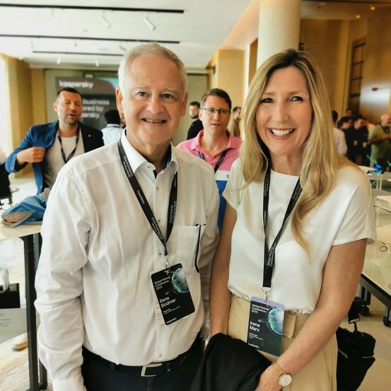 Zwei Personen, ein Mann und eine Frau, stehen lächelnd für ein Foto auf einer Konferenz. Sie tragen weiße Hemden, Lanyards und Ausweise. Im Hintergrund stehen mehrere Personen, die möglicherweise ebenfalls an der Veranstaltung teilnehmen.