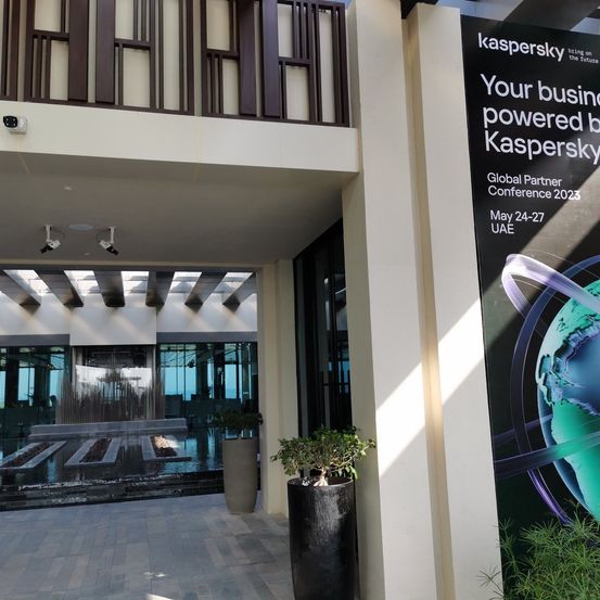 Ein Bild des Außenbereichs eines modernen Gebäudes mit einem Schild, das die Kaspersky Global Partner Conference 2023 ankündigt, mit Datum und Ort. Ein Springbrunnen und Topfpflanzen im Vordergrund.