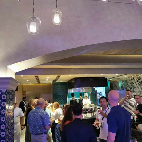 In einem Restaurant stehen mehrere Personen um einen Tresen herum und plaudern möglicherweise. Die Köche sind mit der Essenszubereitung beschäftigt. Das Restaurant hat eine geflieste Decke und Wände.