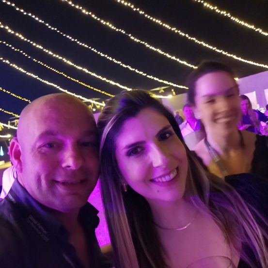 Ein Mann und eine Frau stehen bei Nacht unter einem Baldachin von Lichtern und lächeln für die Kamera. Eine andere Frau ist auch im Hintergrund.