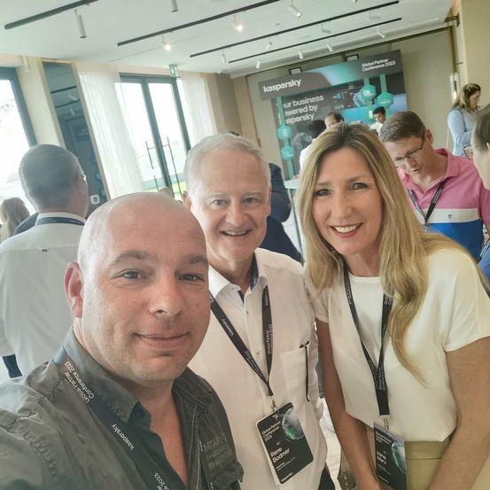 Drei Personen mit Ausweisen lächeln für ein Foto auf einer Konferenz, mit einem Banner hinter ihnen, das 'kaspersky' steht.