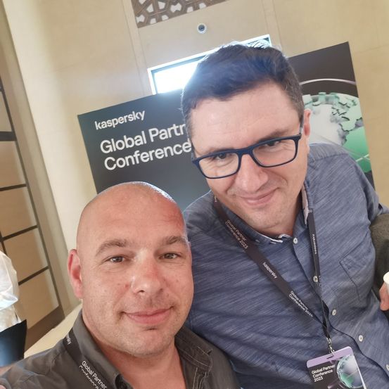 Zwei Männer lächeln für ein Foto auf der Kaspersky Global Partner Conference 2023. Beide tragen Konferenzausweise, und ein Schild hinter ihnen trägt den Text 'Kaspersky Global Partner Conference'.