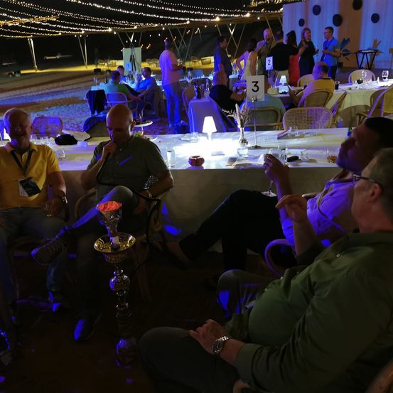Männer sitzen um Tische herum in einer Party-Atmosphäre, einer hält ein Mikrofon. Andere genießen Getränke. Die Szene ist mit warmen Lichtern beleuchtet.
