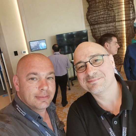 Zwei Männer machen ein Selfie in einem Konferenzraum. Dahinter geht ein Mann an einem Monitor und einer Skulptur vorbei. Ein Schild an der Wand sagt 'Ihr Geschäft, das von Kaspersky angetrieben wird.'