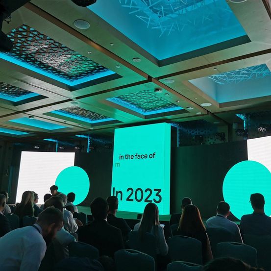 Ein Konferenzraum mit einer großen Projektionsfläche, die 'In der Face von m Im Jahr 2023' anzeigt. Mehrere Teilnehmer sitzen in Reihen, einige unterhalten sich. Der Raum ist gut beleuchtet mit einer Mischung aus Decken- und Umgebungsbeleuchtung.