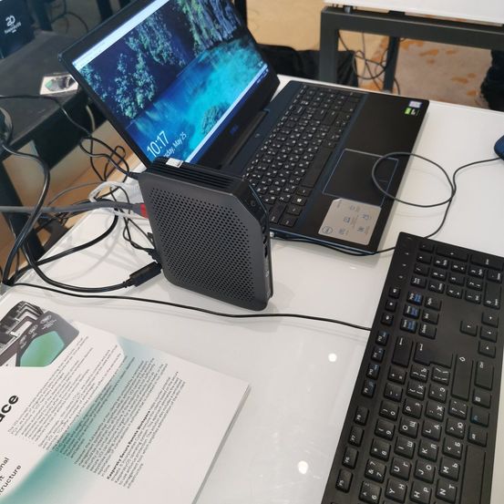 Ein schwarzer Laptop steht auf einem Tisch mit Tastatur und einer schwarzen Box mit USB-Kabel. Der Bildschirm des Laptops zeigt ein Datum. Ein Papier mit Text liegt auf dem Tisch.