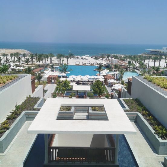 Luftaufnahme eines Resorts mit einem großen Pool, umgeben von Palmen, Liegestühlen und einem Strand mit dem Ozean im Hintergrund.