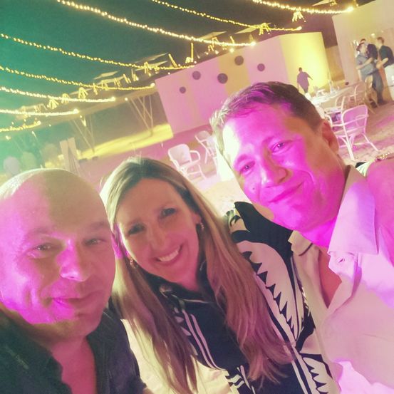 Drei Menschen posieren für ein Selfie auf einer Party. Sie lächeln unter rosa Lichtern mit Lichterketten darüber. Hinter ihnen ist eine rosa Wand und eine Tanzfläche.