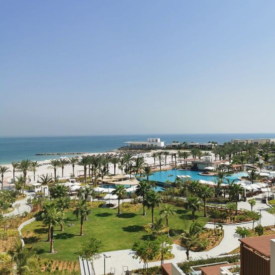 Luftaufnahme eines Resorts mit Strand, Palmen und einem großen Pool. In der Ferne sind Gebäude zu sehen. Der Himmel ist klar.
