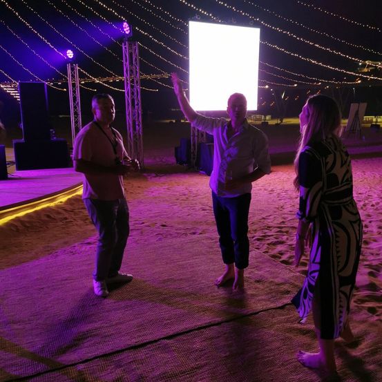 Drei Menschen stehen bei Nacht auf einem sandigen Strand, beleuchtet von lila Lichtern. Ein Mann zeigt auf eine große Leinwand, während zwei andere zuschauen.