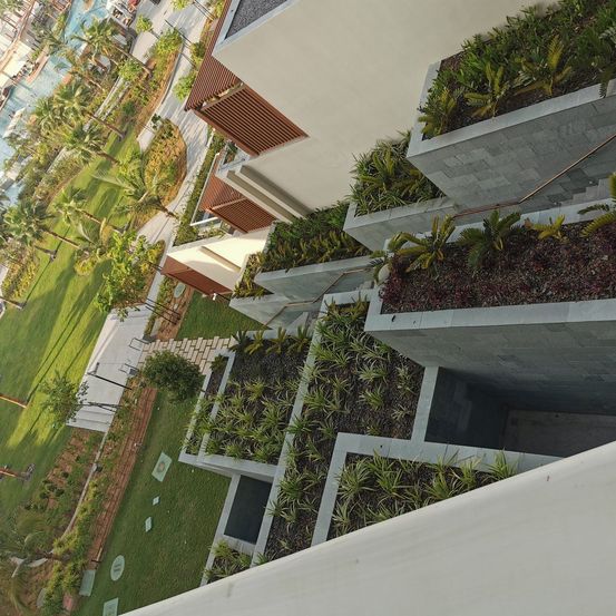 Eine Draufsicht auf ein modernes Gebäude mit vertikalen Gärten. Das Gebäude hat ein einzigartiges Design mit Betonkästen, die Grünpflanzen enthalten. Die Umgebung ist von üppigem Gras und Palmen umgeben.