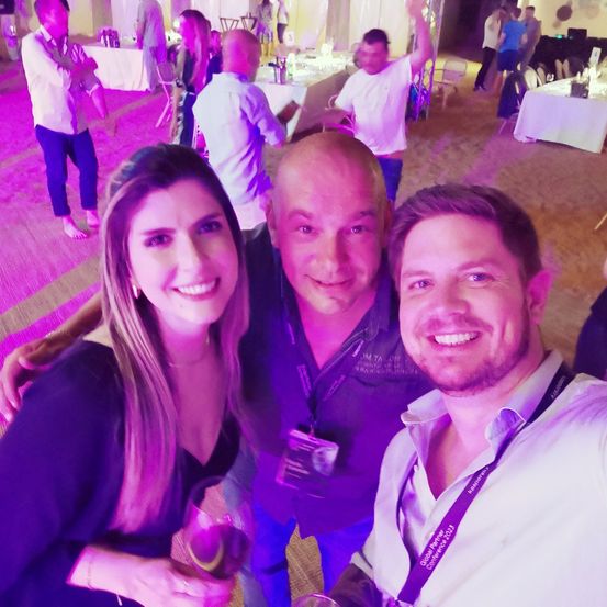 Eine lächelnde Frau und zwei Männer posieren für ein Foto auf einer Party mit lila Beleuchtung und halten Getränke in der Hand. Sie tragen Namensschilder. Im Hintergrund sind Leute an Tischen versammelt, und ein Mann steht auf einem Stuhl.
