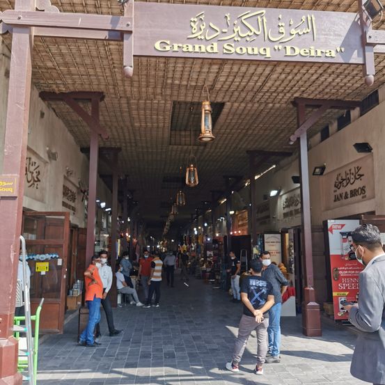Der Grand Souq Deira in Dubai, mit Menschen, die Masken tragen, Geschäften mit arabischen Beschilderungen und einer Laterne, die von der Decke hängt.