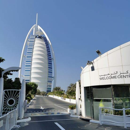 Ein modernes Gebäude mit arabischem Text an seiner Seite. Der Eingang hat ein Schild mit der Aufschrift 'Welcome Center'. Es gibt eine Straße mit einer weißen Linie und einem weißen Tor.