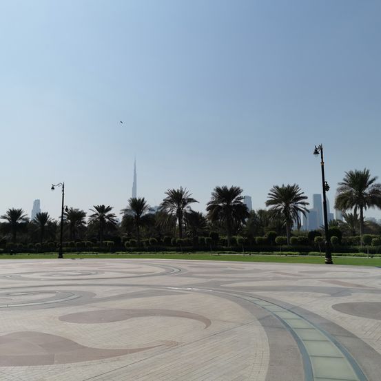 Ein großer kreisförmiger Park mit einem Muster auf dem Boden, umgeben von Palmen, Straßenlaternen und Büschen. In der Ferne stehen hohe Gebäude.