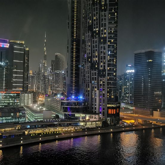 Ein nächtlicher Blick auf eine Stadtsilhouette mit zahlreichen Hochhäusern, die mit verschiedenen Lichtern beleuchtet sind, und einem Gewässer, das die Stadtlichter reflektiert.